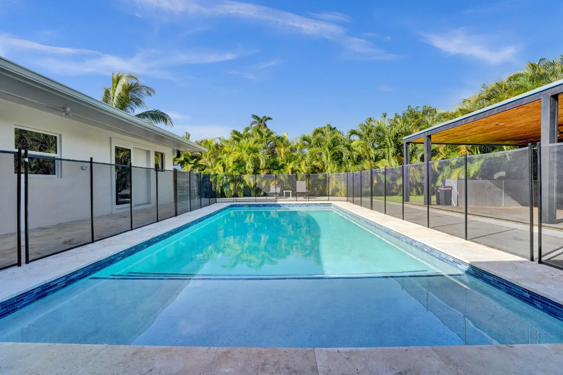 Rectangular blue pool with travertine deck, safety fence, and palm-lined backyard