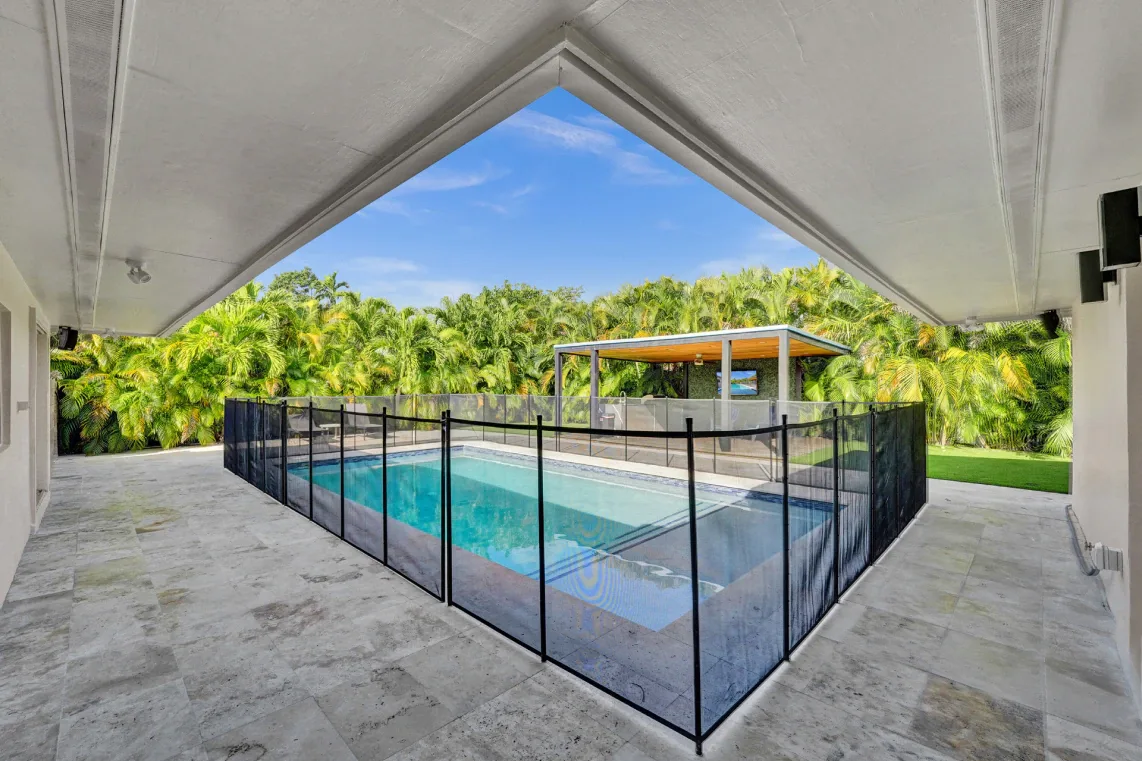 Covered stone patio overlooking a fenced rectangular pool and lush tropical landscaping